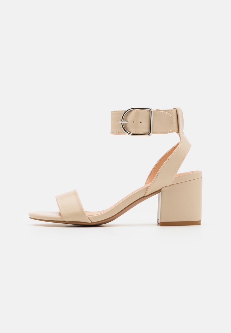 Sandale beige à bout ouvert avec talon carré et large bride à la cheville avec une grande boucle argentée sur fond blanc.