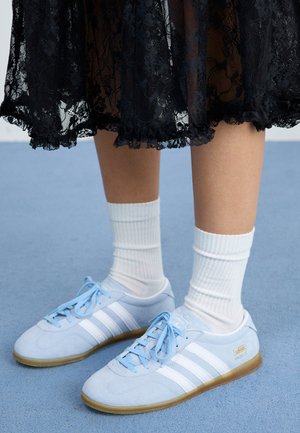 Light blue sneakers with white stripes and white socks, worn with a black lace skirt, on a blue carpeted surface.