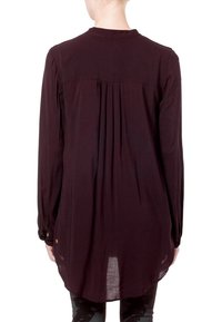 Blouse à manches longues de couleur bordeaux foncé avec un détail de pli au dos, ourlet arrondi et poignets à boutons, fabriquée dans un tissu léger et fluide.