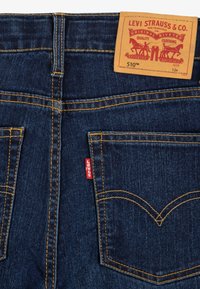 Mörkblå Levi's jeans bakvy som visar fickorna, orange sömnad, röd Levi's-tag och brun läderpatch med varumärkes- och storleksinformation.