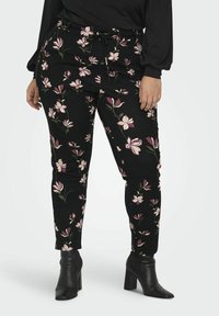 Naine kannab musti slim-fit pükse roosa ja valge lillekirjaga, musta pikkade käistega särk ja musti kontsaga ankle-bote.