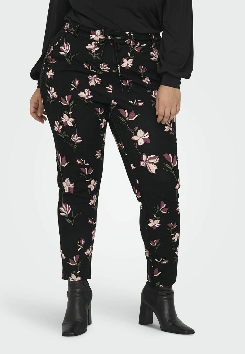 Naine kannab musti slim-fit pükse roosa ja valge lillekirjaga, musta pikkade käistega särk ja musti kontsaga ankle-bote.