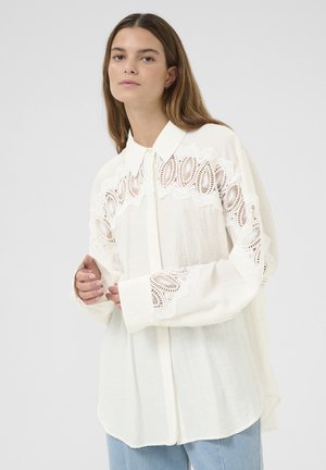 Camicia bianca con bottoni, caratterizzata da dettagli in pizzo intricati sulle maniche, texture morbida e vestibilità rilassata, completata da un colletto classico.