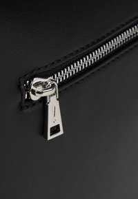 Roh černé kožené tašky s kovovým zipem a obdélníkovým táhlem označeným detailem výkřičníku. Hladký povrch.