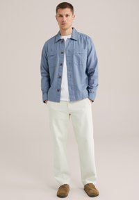 Camisa de botones azul claro con dos bolsillos en el pecho, combinada con pantalones blancos y zapatos marrones sin cordones, con una textura suave.