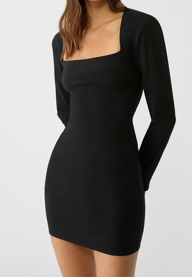 Femme portant une robe mini noire ajustée à manches longues et à encolure carrée, se tenant devant un fond clair uni.