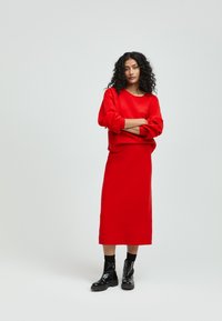 Roter Pullover und passender Midirock, beide aus einem strukturierten Stoff. Das Model trägt schwarze Knöchelschäfte und steht mit verschränkten Armen.