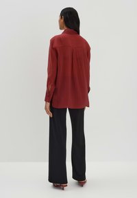 Camicia rossa a maniche lunghe con vestibilità comoda, dettaglio della piega posteriore e aperture ai polsini, abbinata a pantaloni neri a gamba larga e scarpe con tacco.