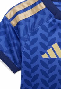 Manga de camiseta deportiva azul con un patrón de hojas en azul oscuro, franjas doradas en los hombros, puño acanalado y parte del logo dorado de Adidas en el pecho.