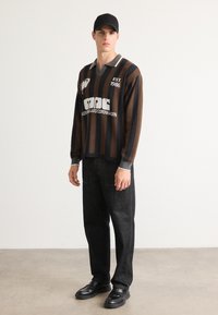 Giovane uomo che indossa un cappello nero, una camicia a maniche lunghe a strisce marroni e nere con del testo, pantaloni neri e scarpe nere mentre sta in piedi su un pavimento grigio.