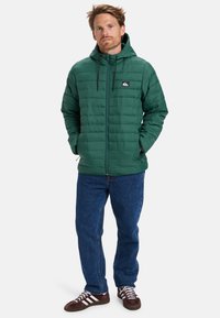 Grüne Daunenjacke mit Kapuze, Reißverschluss vorne und einem Logo-Patch auf der Brust. Kombiniert mit blauen Jeans und braunen Schuhen mit weißen Streifen.