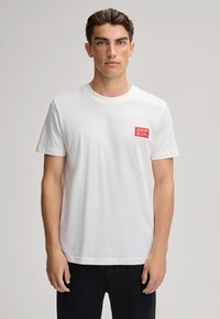 Biała bawełniana koszulka z okrągłym dekoltem i krótkimi rękawami. Posiada czerwony kwadratowy logo na lewej piersi. Prosty fason, luźny krój.