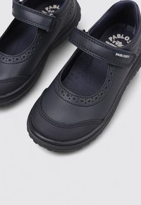 Par de zapatos para niños estilo Mary Jane en color negro con detalles perforados, correas de Velcro y suelas de goma duraderas sobre un fondo claro.