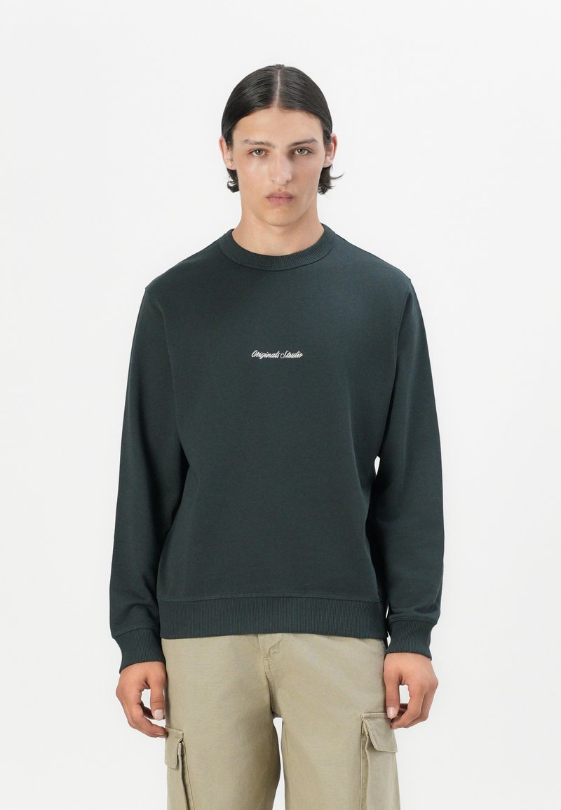 Sudadera de color verde oscuro hecha de una tela suave, con un cuello y puños acanalados, y un pequeño logo blanco de "Original Studio" en el pecho.