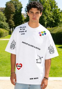 Jeune homme aux cheveux bouclés et foncés portant un T-shirt blanc oversize avec différents textes et symboles imprimés en noir et en couleur en extérieur dans un parc.