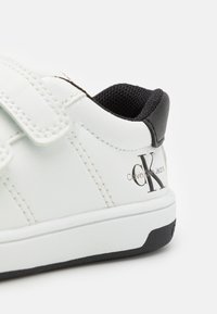 Vit skinnsneaker med svarta detaljer, utrustad med kardborreband och ett texturerat svart innerfoder. "Calvin Klein Jeans"-logotyp nära hälen.