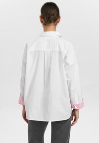 Camicia bianca con colletto, maniche lunghe, con polsini a righe rosa e piega sul retro. Il tessuto sembra liscio e leggero.