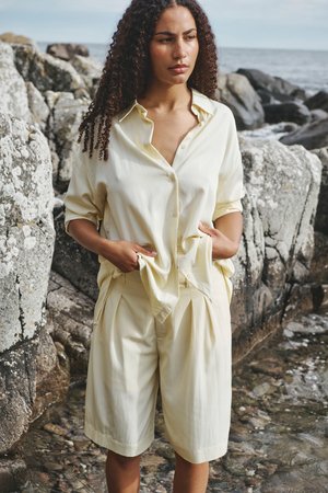 Mujer con cabello rizado, camisa amarilla clara abotonada y pantalones cortos, de pie junto a la costa rocosa, mirando pensativamente hacia un lado.