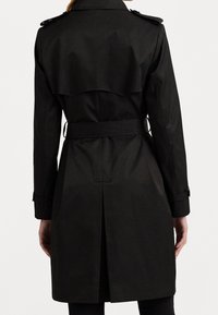 Svart trenchcoat med bälte i midjan, axelklaffar och ett lager-på-lager-design på bakstycket. Slät material med en något glänsande yta.