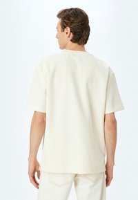 Crème katoenen t-shirt met korte mouwen, ronde hals, relaxed fit en subtiele textuur, gezien van de achterkant, gecombineerd met lichte broek.