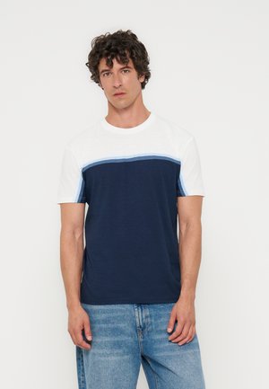 Uomo con capelli ricci che indossa una t-shirt bianca e blu navy a blocchi di colore e jeans azzurri, in piedi davanti a uno sfondo bianco semplice.