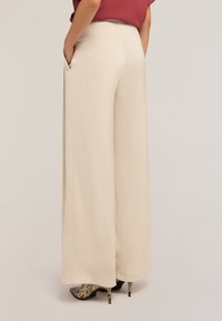 Pantalons larges beiges avec une texture lisse, dotés de poches latérales et d'une taille haute, associés à des chaussures à talons à motifs.