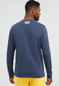 Mann, der ein blaues Langarmhemd mit einem weißen Etikett am oberen Rücken und gelbe Hosen trägt, von hinten zu sehen.