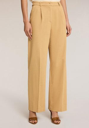 Lichtbeige, wijde broek met een gestructureerde afwerking, voorzien van voorplooien en zijzakken. Gecombineerd met bruine sandalen met open tenen.