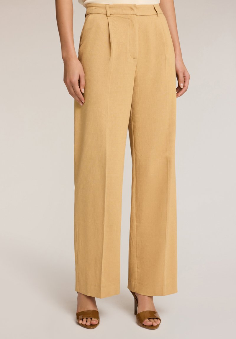 Lichtbeige, wijde broek met een gestructureerde afwerking, voorzien van voorplooien en zijzakken. Gecombineerd met bruine sandalen met open tenen.