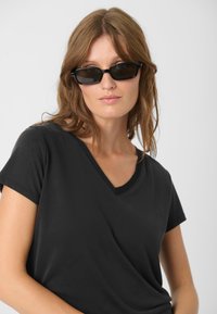 Černé tričko s výstřihem do V vyrobené z měkké tkaniny, s krátkými rukávy. Model má na sobě oversized černé sluneční brýle, které zvýrazňují moderní vzhled outfitu.