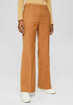 Vrouw draagt roestkleurige broek met hoge taille en wijde pijpen, gecombineerd met witte sneakers en een beige top met lange mouwen.