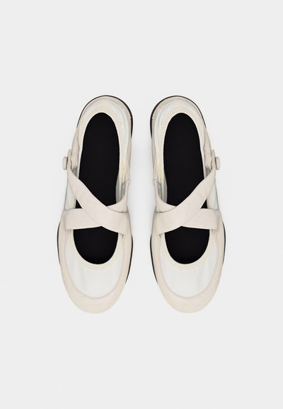 Ζευγάρι μπεζ παπούτσια Mary Jane με στρογγυλές μύτες, μαύρα πάτακια και σταυρωτά λουράκια με κουμπιά κλεισίματος σε λευκό φόντο.