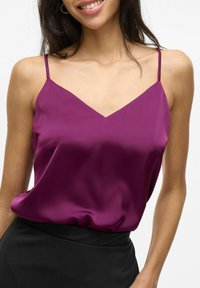 Camisole en satin violet avec un décolleté en V et des bretelles fines, offrant une coupe décontractée, associée à un vêtement noir. Texture lisse.