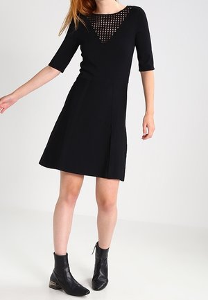 Femme portant une robe noire longueur genoux avec des manches mi-longues et un détail en mesh avec un col en triangle, associée à des bottines noires.