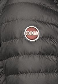 En pufferjacka i svart tyg med quiltad textur, som har en cirkulär brun och vit Colmar-logotyp i paddning på övre vänstra sidan.