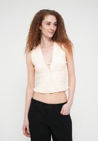 Νεαρή γυναίκα με καστανέρυθρα σγουρά μαλλιά, φορώντας αμάνικο εκρού τοπ με ρυτίδες και μαύρο παντελόνι, στέκεται μπροστά σε απλή λευκή φόντο.