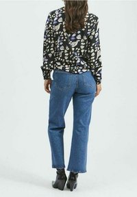 Frau mit langen braunen Haaren trägt eine schwarze Blumenbluse, blaue gerade Jeans und schwarze, blockierte Knöchelstiefel und steht vor einem schlichten Hintergrund.