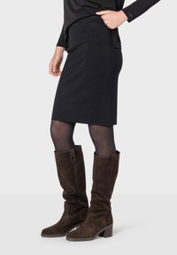 Černá sukně ke kolenům vyrobená z pletené tkaniny, v kombinaci s dlouhými hnědými semišovými botami s blokovým podpatkem a moderními designovými akcenty.