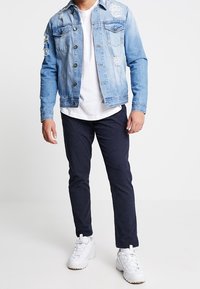 Man in een lichtblauwe distressed denim jack, witte t-shirt, donkere gestreepte broek en witte sneakers, staand tegen een effen achtergrond.
