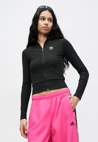 Fekete cipzáros rövid felső bordázott textúrával és egy kis fehér logóval; élénk rózsaszín melegítőnadrággal párosítva, fekete oldalsávokkal.