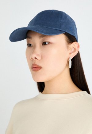 Blå bomullslue med buet skjerm. Designet inkluderer en knapp på toppen og en logo på siden, kombinert med en sweatshirt i nøytral farge.