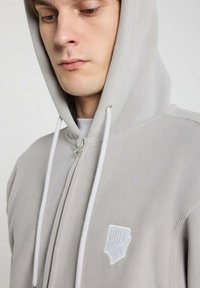 Haut de jogging zippé gris en polaire avec une étiquette de logo blanche, cordons de serrage blancs réglables et une texture douce. Comprend une capuche et des coutures discrètes.