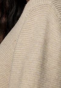 Nahaufnahme eines beigen Strickstoffs auf einem Kleidungsstück, mit dunklen Haaren, die teilweise im Hintergrund sichtbar sind.