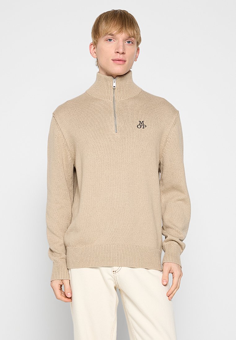 Beige gebreide trui met hoge kraag en halve rits. Ribgebreide mouwen en zoom. Heeft een klein geborduurd logo op de linkerborst.