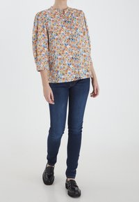Blouse florale bleu clair, ornée de fleurs orange et roses, avec un col rond et des manches trois-quarts. Portée avec un jean skinny bleu foncé.