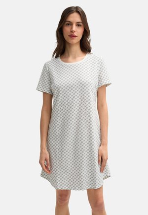 Młoda kobieta stojąca, ubrana w białą sukienkę z krótkim rękawem z delikatnym szarym wzorem kołowym, neutralne tło.