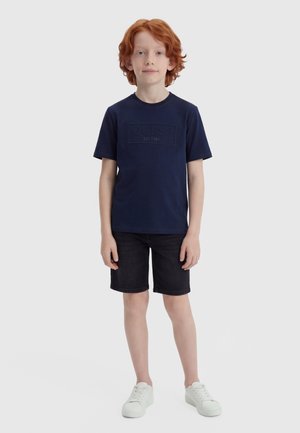 Enfant aux cheveux roux debout, portant un t-shirt bleu marine à manches courtes, un short noir et des baskets blanches sur un fond blanc uni.