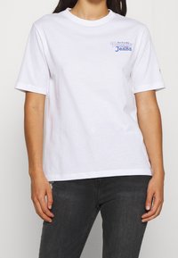 T-shirt en coton blanc à manches courtes, arborant un logo "Tommy Jeans" bleu sur la partie gauche de la poitrine. Couleur unie, coupe décontractée.