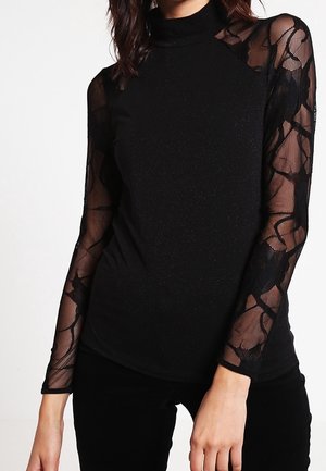 Femme portant un haut noir ajusté avec des manches longues en dentelle florale transparente et un col montant noir uni et un buste.