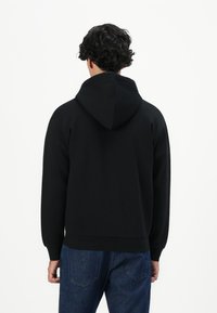Mann med krøllete mørkt hår iført en enkel svart hettegenser og blå jeans, stående med ryggen til mot en hvit bakgrunn.